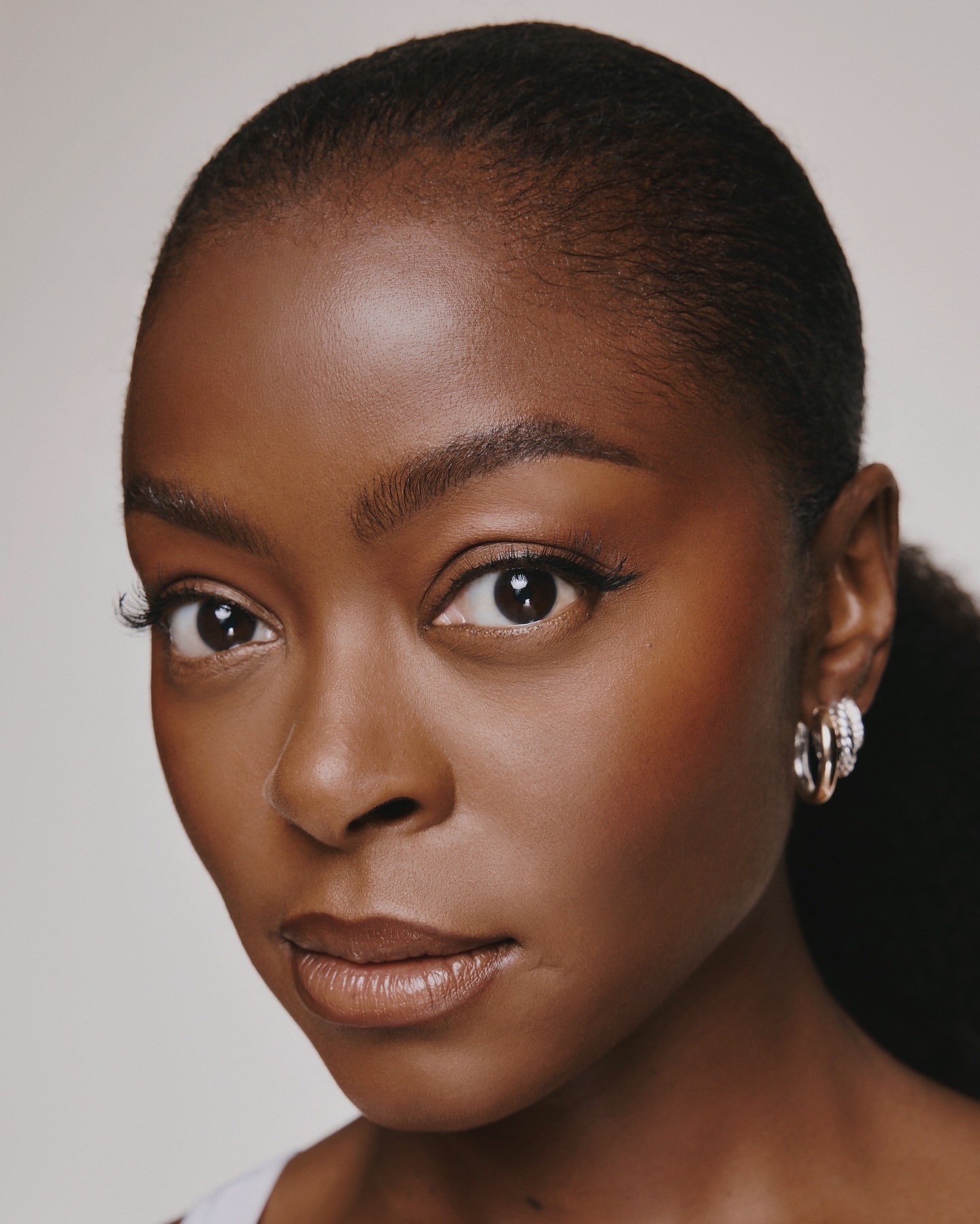 Close-up portrait of a beautiful African American woman with radiant makeup, wearing hoop earrings against a neutral backdrop.