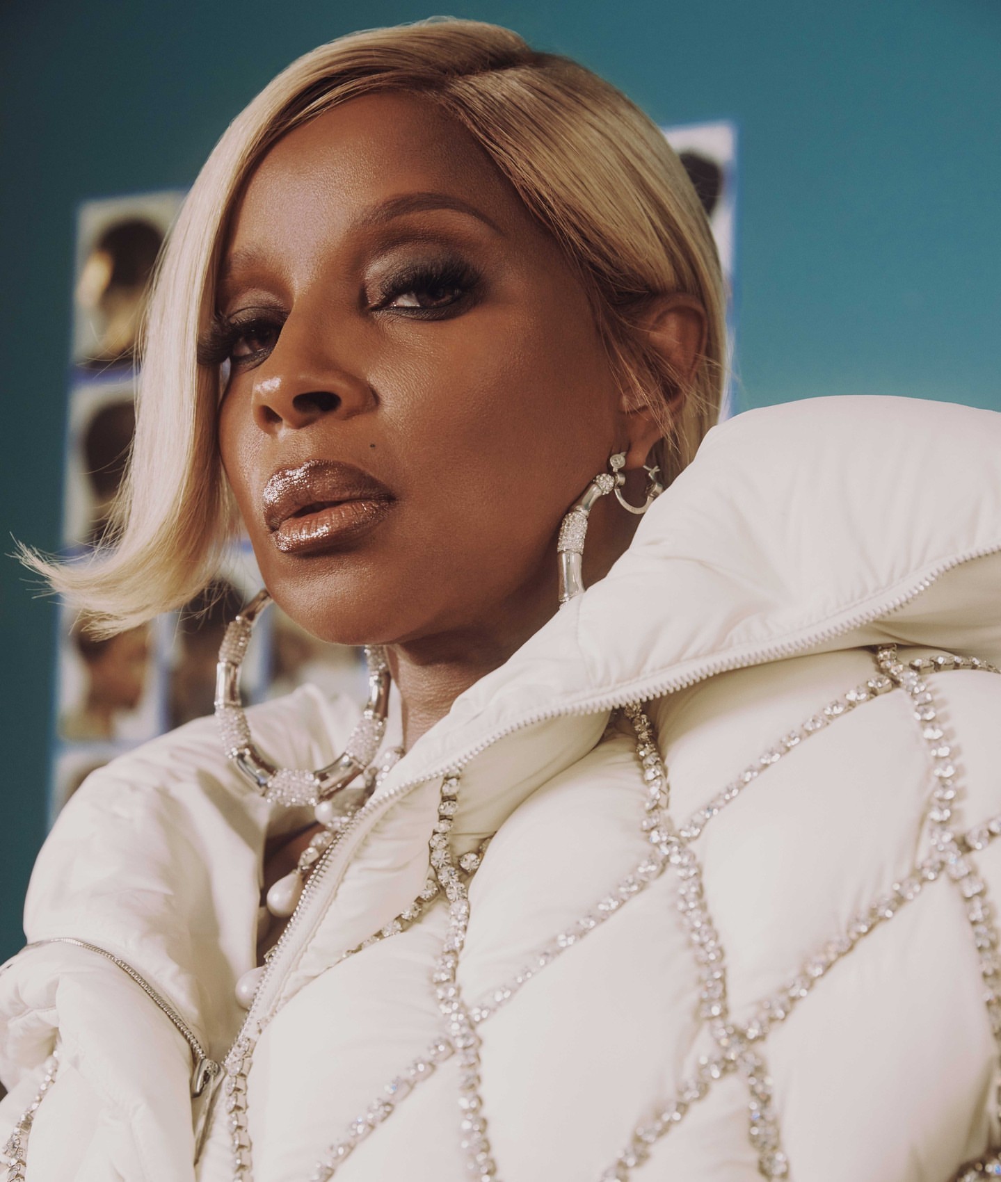 Close-up portrait of a woman with blonde hair wearing a white puffer jacket and hoop earrings against a blue background.