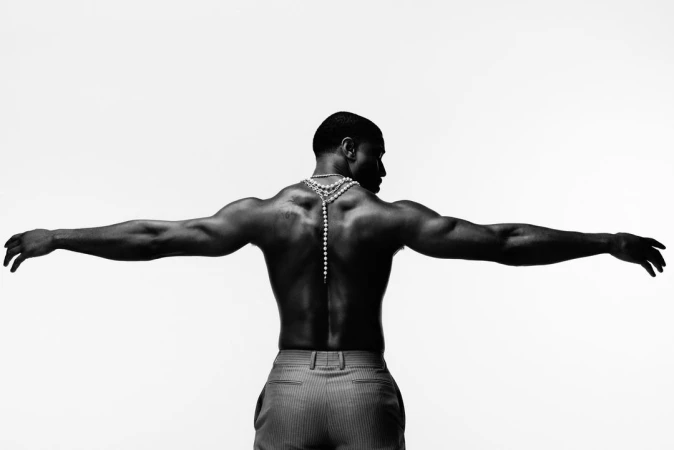 Shirtless man with arms outstretched wearing layered pearl necklaces viewed from behind against white background