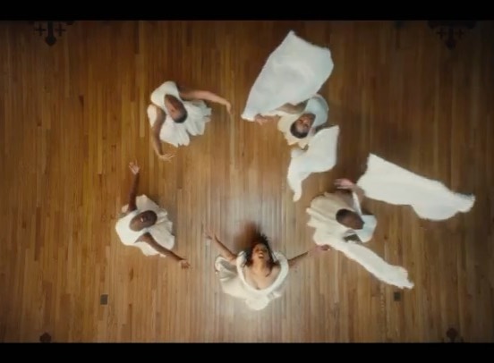 Overhead shot of six people in white dresses in a circle on a light brown wood floor, with arms raised and outstretched.