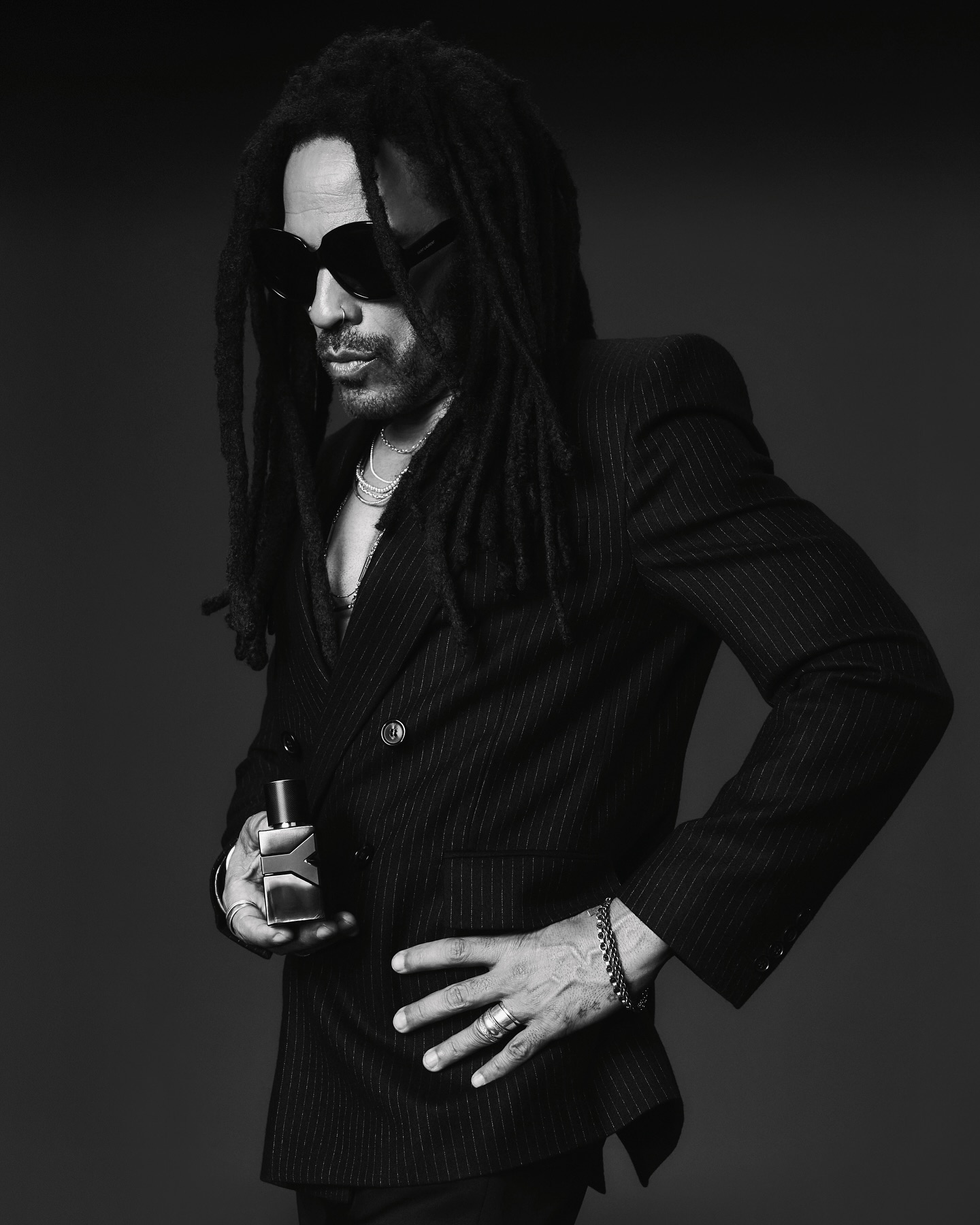Lenny Kravitz wearing a dark pinstripe blazer and sunglasses holding a fragrance bottle in a portrait by Adrienne Raquel