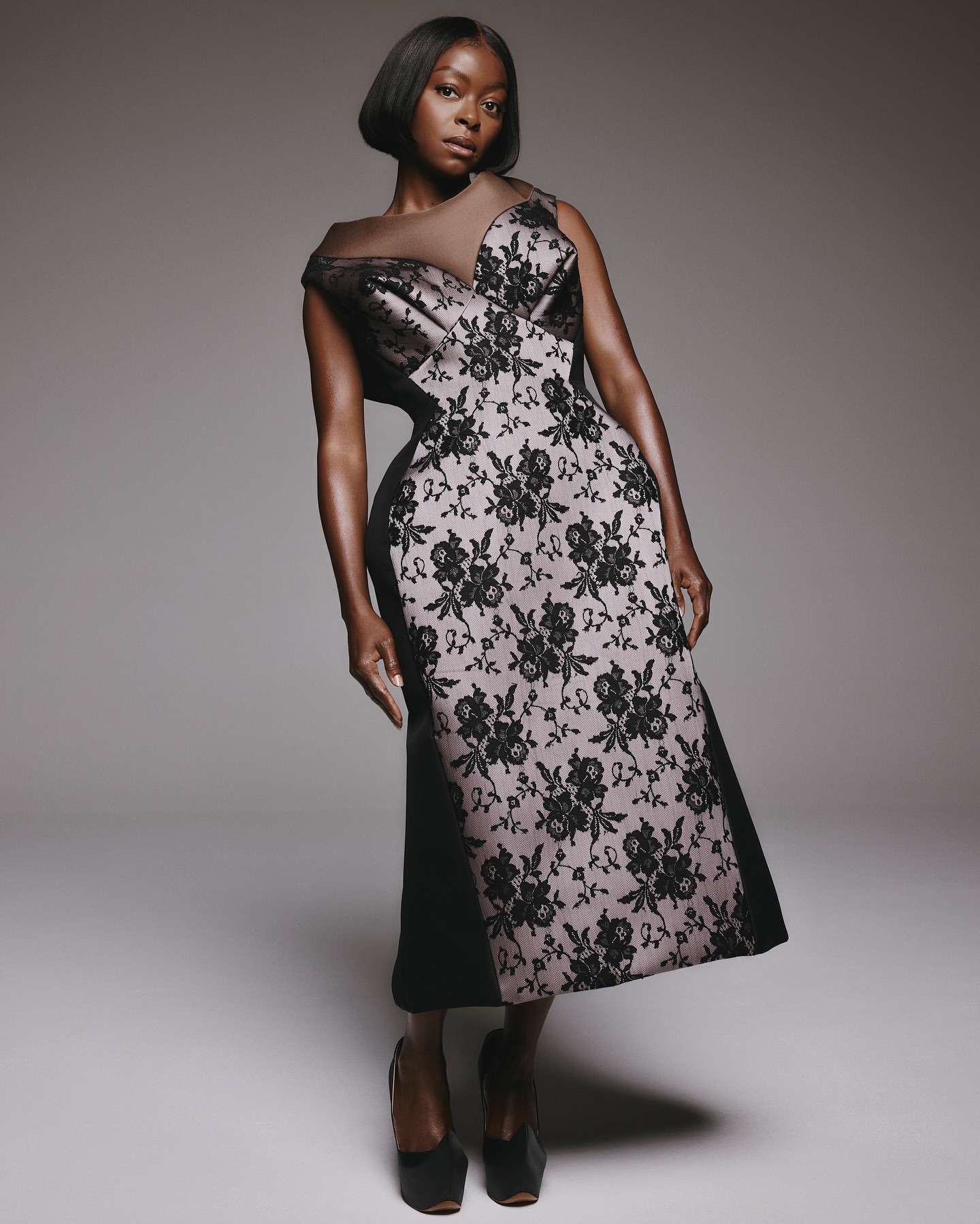 Elegant woman in a black and floral dress posing in a studio. She has a short bob haircut and wears dark heels.