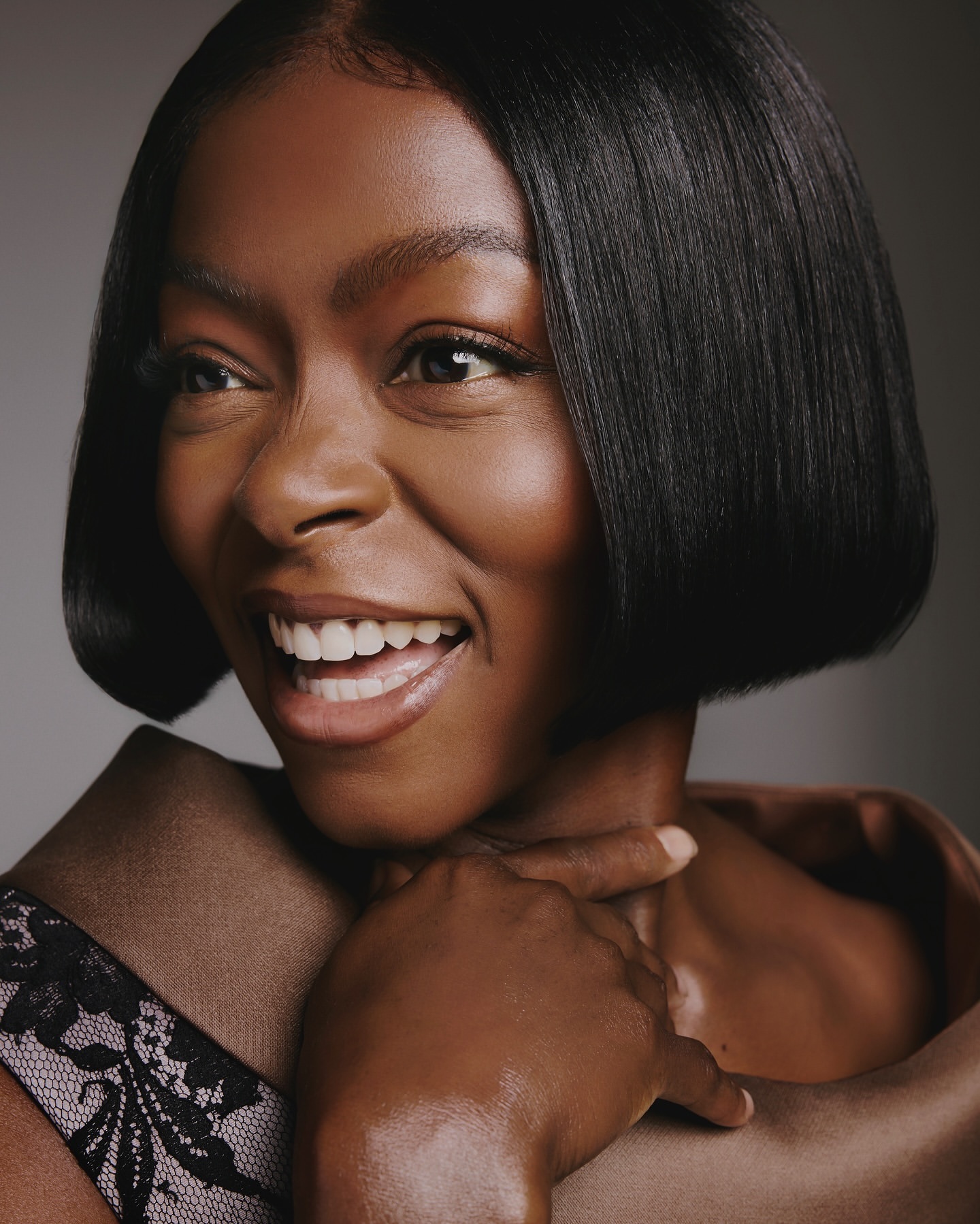 Portrait of a smiling Black woman with a bob hairstyle and natural makeup is embraced by a person with lacy-trimmed clothing.