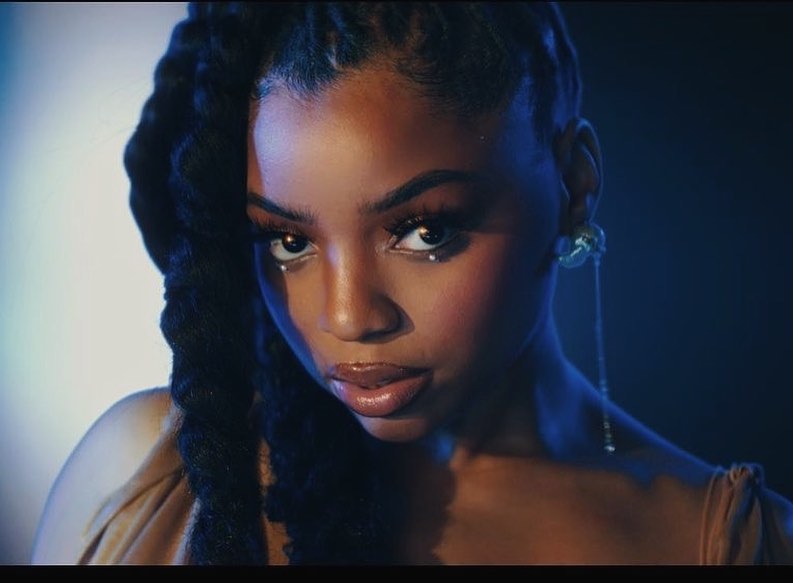 Close-up portrait of an African-American woman with dark braided hair, wearing an off-the-shoulder tan dress, against a blue background.