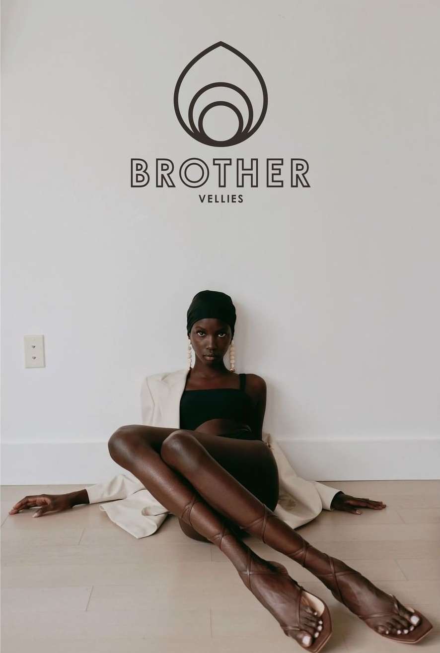 Fashion shot of a Black female model wearing a headscarf, dress, blazer, and strappy high-heeled sandals, sitting on the floor in a studio.