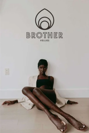 Fashion shot of a Black female model wearing a headscarf, dress, blazer, and strappy high-heeled sandals, sitting on the floor in a studio.