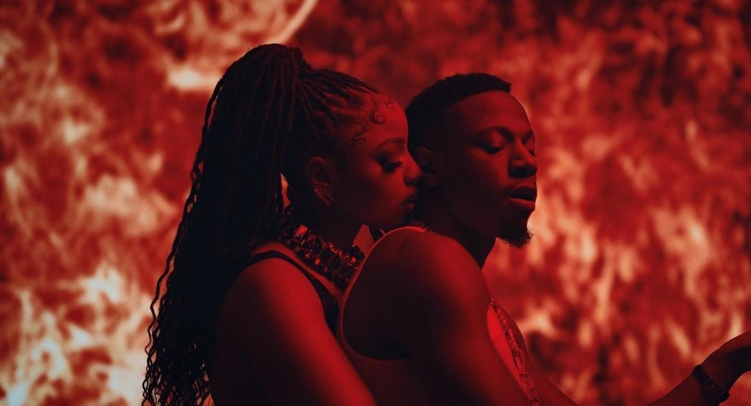 Two adult figures, a Black woman with braided hair and a Black man, pose closely against a fiery red backdrop with a music video aesthetic.
