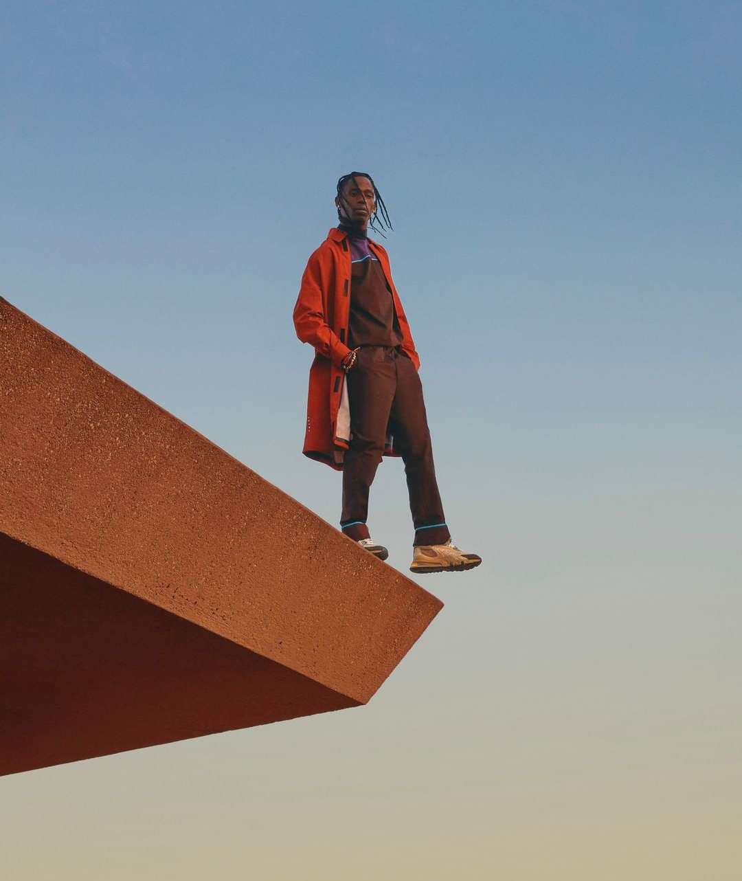 A male adult model stands on a concrete geometric structure against a blue gradient sky wearing brown pants and a red coat.