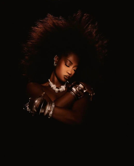 Portrait of an adult woman with a voluminous afro hairstyle, wearing a white necklace and silver bracelets against a dark background.