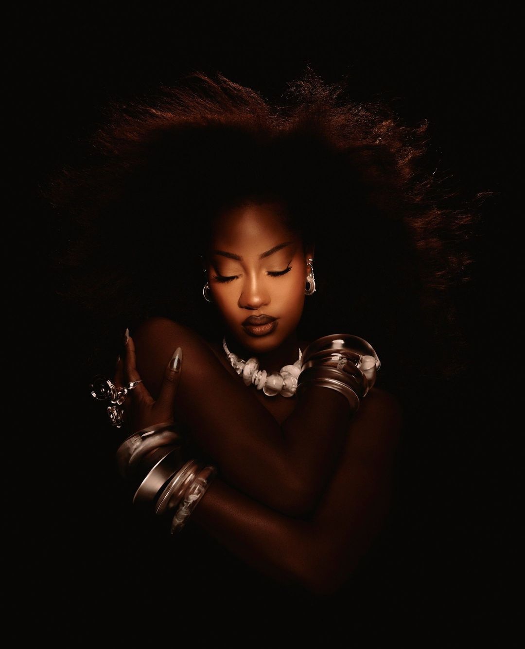 Dark-skinned adult woman with afro embracing herself, wearing bracelets and necklace in a dark photo studio setting with moody lighting.