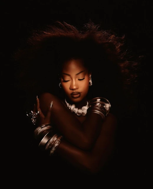 Dark-skinned adult woman with afro embracing herself, wearing bracelets and necklace in a dark photo studio setting with moody lighting.