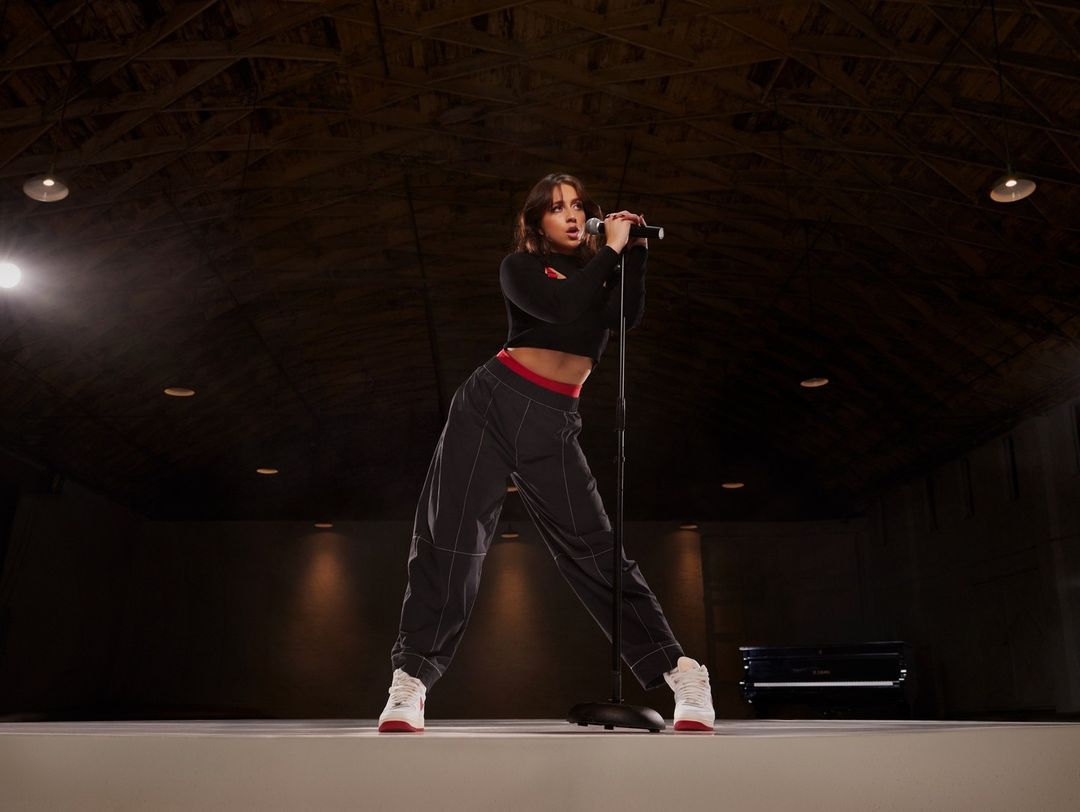 A woman sings into a microphone on stage in a dark venue wearing a black crop top, grey pants and white sneakers with red accents.