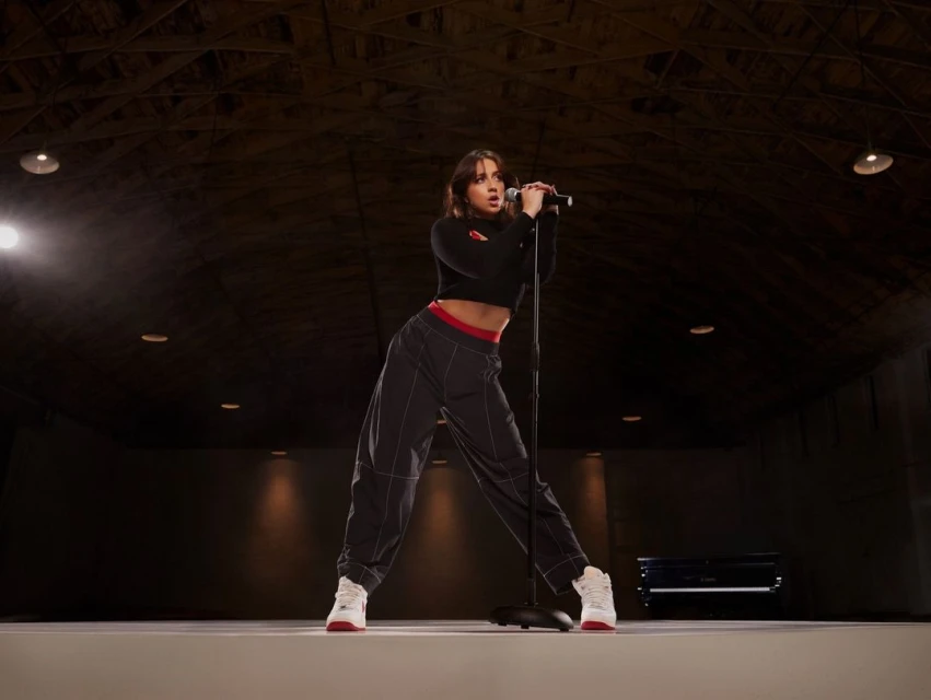 A woman sings into a microphone on stage in a dark venue wearing a black crop top, grey pants and white sneakers with red accents.