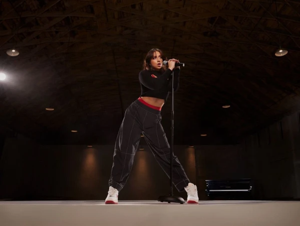A woman sings into a microphone on stage in a dark venue wearing a black crop top, grey pants and white sneakers with red accents.