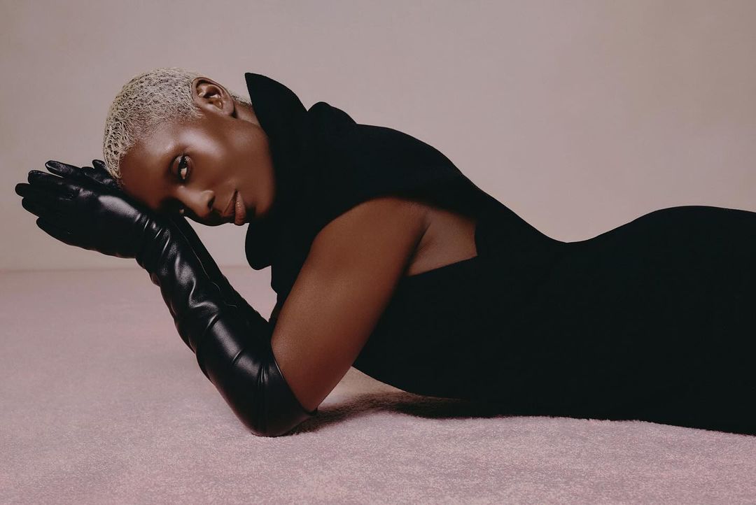An adult female model with short blonde hair poses in a black dress and long black gloves against a light pink seamless background.