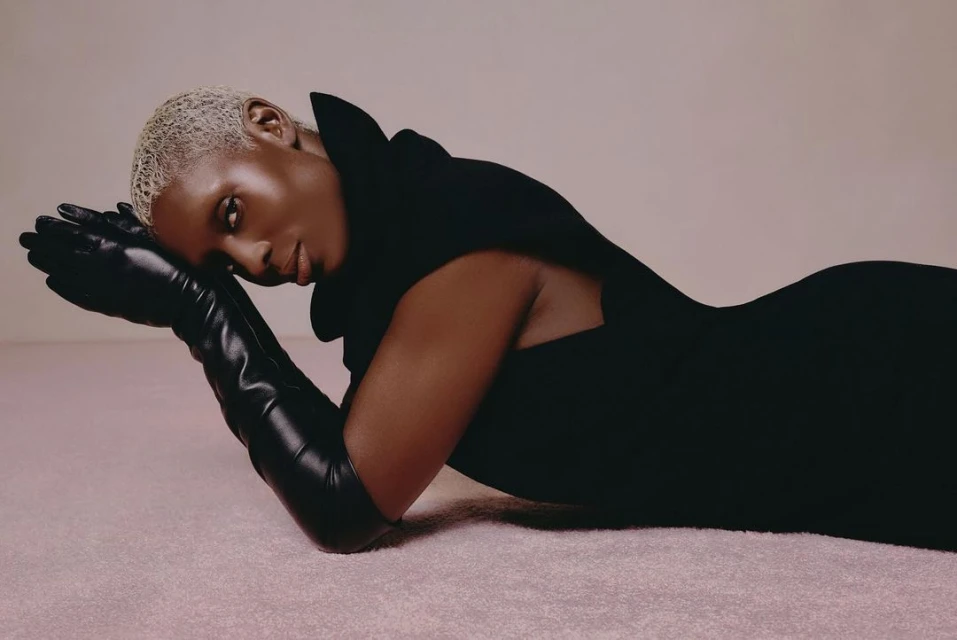 An adult female model with short blonde hair poses in a black dress and long black gloves against a light pink seamless background.