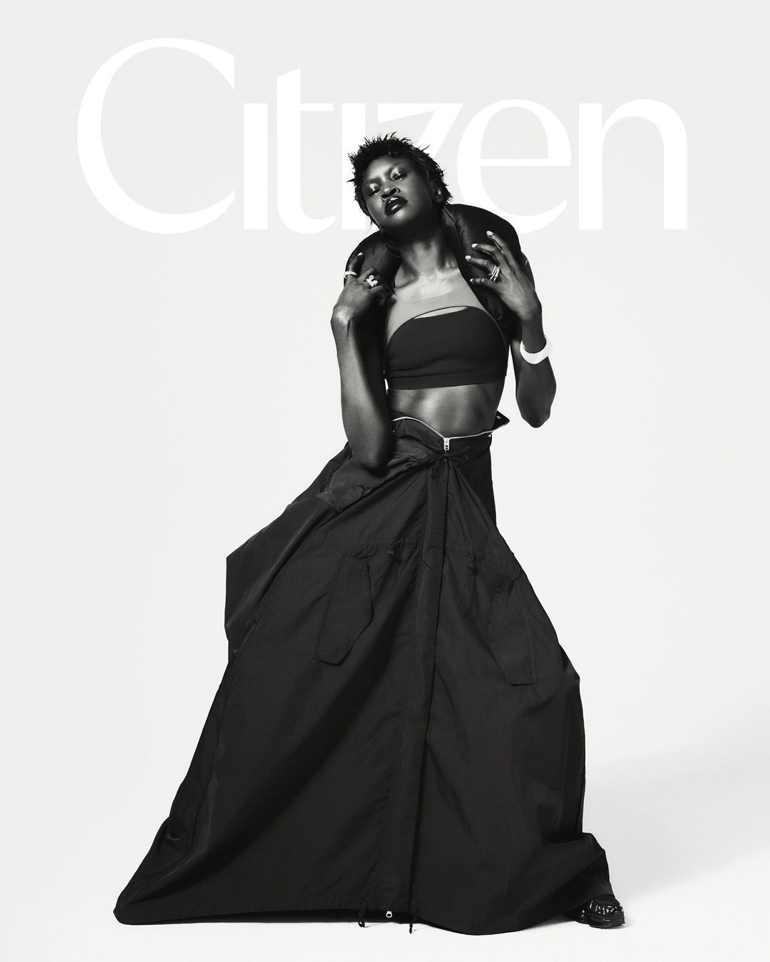 Monochrome fashion portrait of Black female model in crop top, long skirt, and leather jacket against a neutral backdrop.