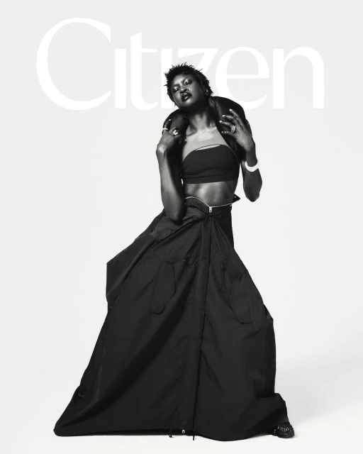 Monochrome fashion portrait of Black female model in crop top, long skirt, and leather jacket against a neutral backdrop.