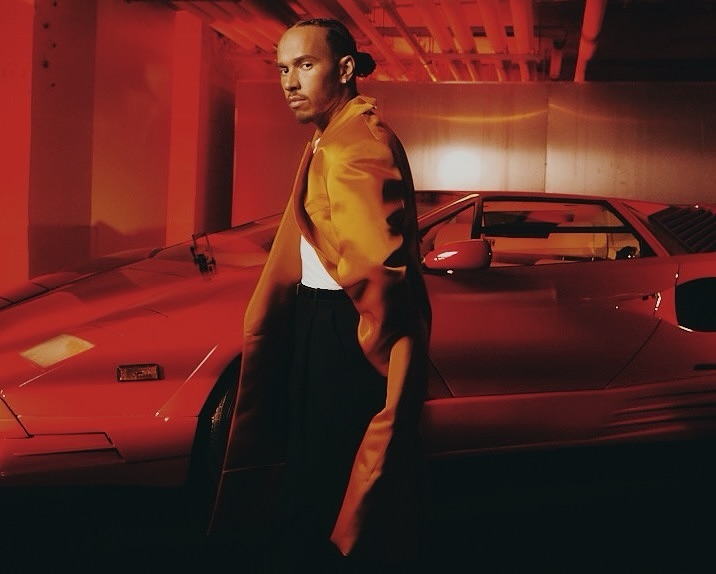 Adult male in a gold satin jacket poses with a red sports car indoors in a dimly lit environment with red ambient lighting.