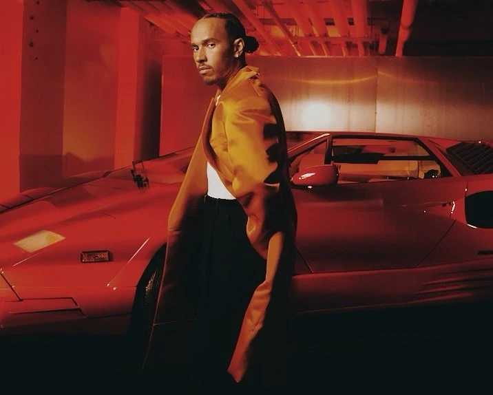 Adult male in a gold satin jacket poses with a red sports car indoors in a dimly lit environment with red ambient lighting.