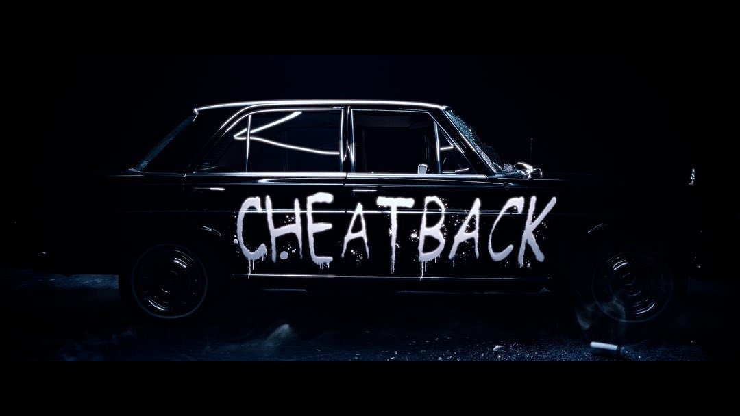 Vintage black car with graffiti-style 'CHEATBACK' writing along the side, parked indoors in a dimly lit environment with a dramatic spotlight.