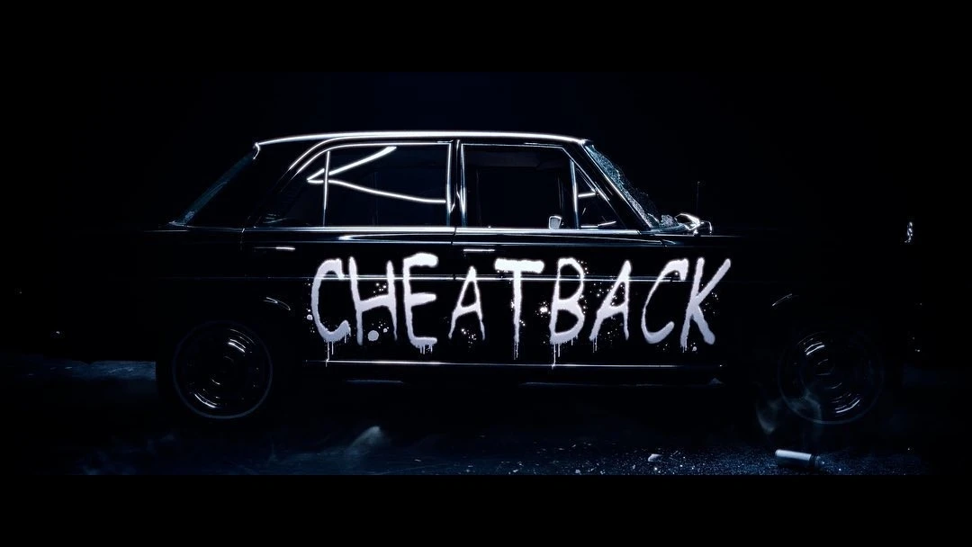 Vintage black car with graffiti-style 'CHEATBACK' writing along the side, parked indoors in a dimly lit environment with a dramatic spotlight.