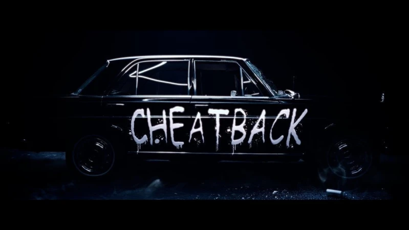 Vintage black car with graffiti-style 'CHEATBACK' writing along the side, parked indoors in a dimly lit environment with a dramatic spotlight.
