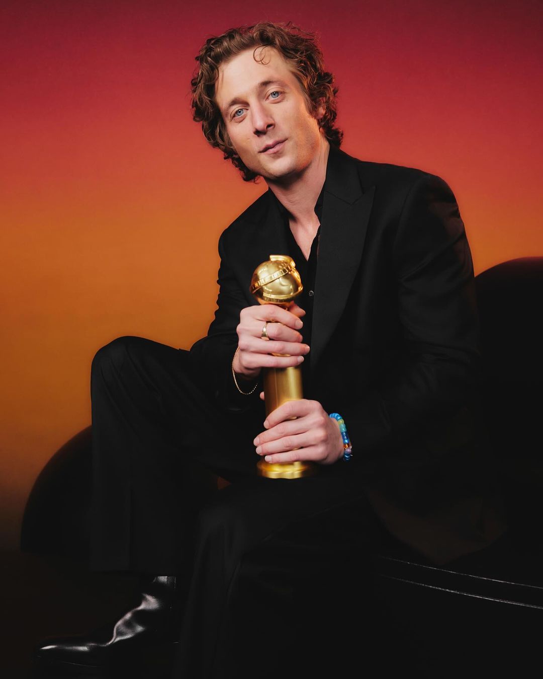 Portrait of a curly-haired man in a black suit holding a gold award against a gradient orange and red background in a studio.