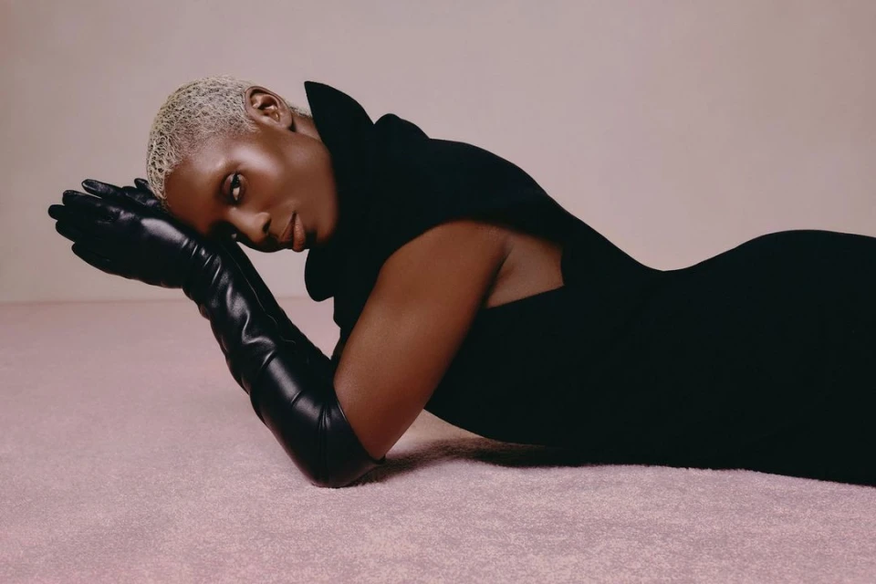 A woman with short blonde hair lies on her side wearing a black dress and elbow-length black gloves against a light pink background.