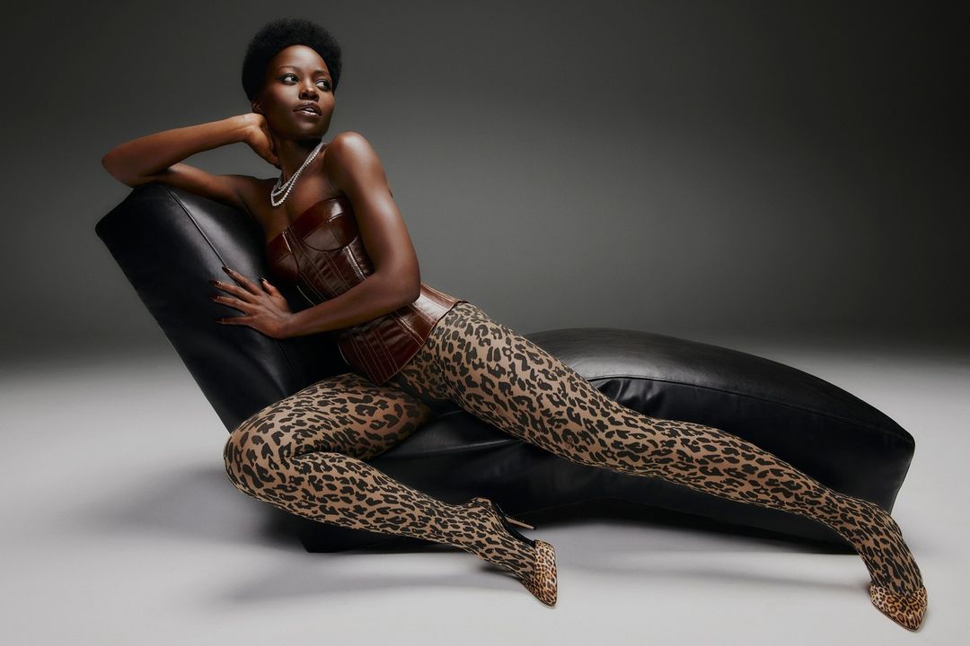 A woman with dark skin and short hair is reclining on a black chaise lounge wearing a brown corset and leopard print tights and heels.