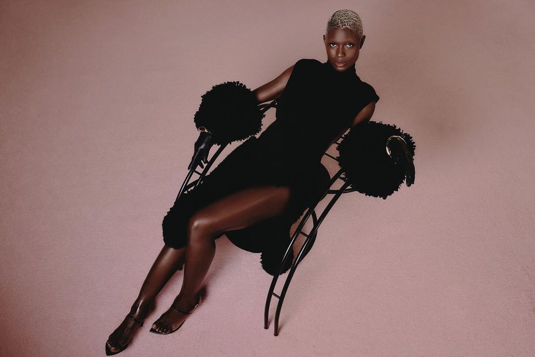 A person with short bleached hair, wearing a fitted black dress, sits on a modern chair with black fuzzy armrests against a pink background.