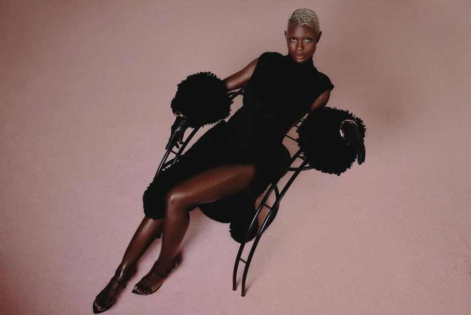 A person with short bleached hair, wearing a fitted black dress, sits on a modern chair with black fuzzy armrests against a pink background.
