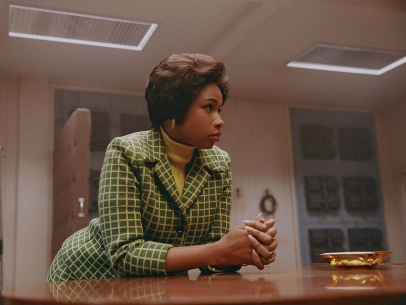 Woman with dark hair in green and yellow patterned jacket sitting at a table in an indoor environment, looking away from the camera.