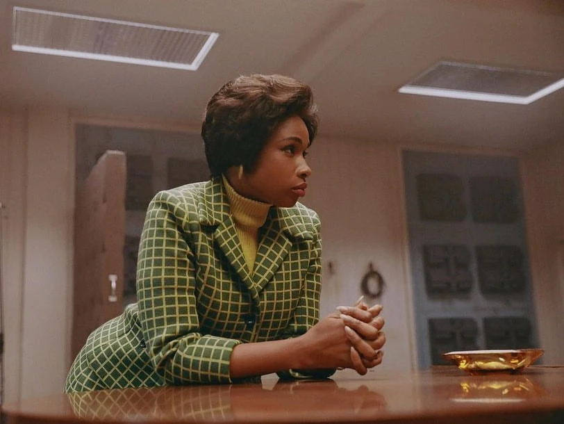 Woman with dark hair in green and yellow patterned jacket sitting at a table in an indoor environment, looking away from the camera.