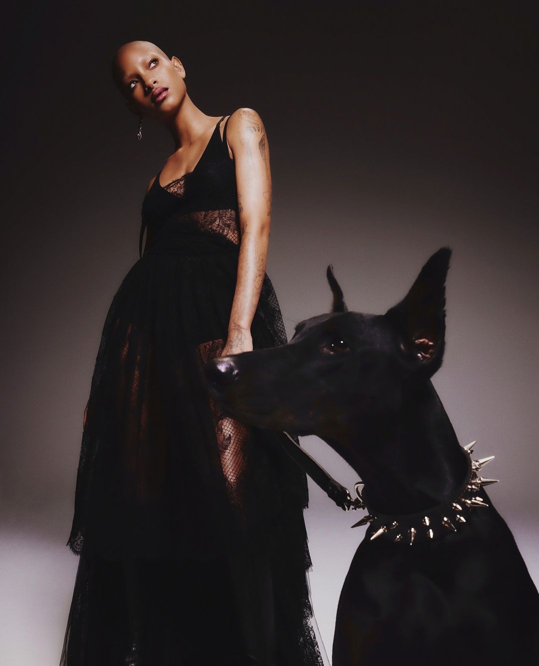 Adult female in a dark dress poses with a black Doberman wearing a spiked collar against a neutral studio backdrop.