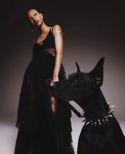 Adult female in a dark dress poses with a black Doberman wearing a spiked collar against a neutral studio backdrop.