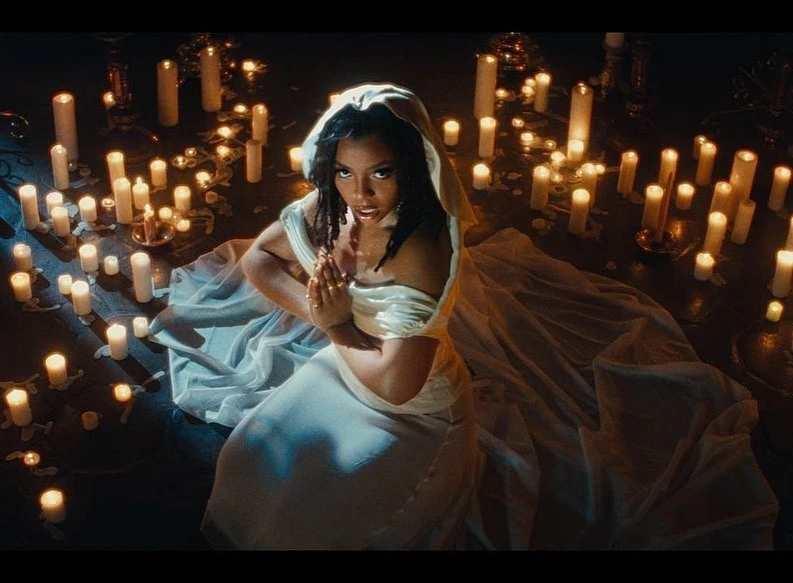 Woman in white dress kneeling surrounded by lit candles, creating a warm and ethereal atmosphere in a dimly lit space.