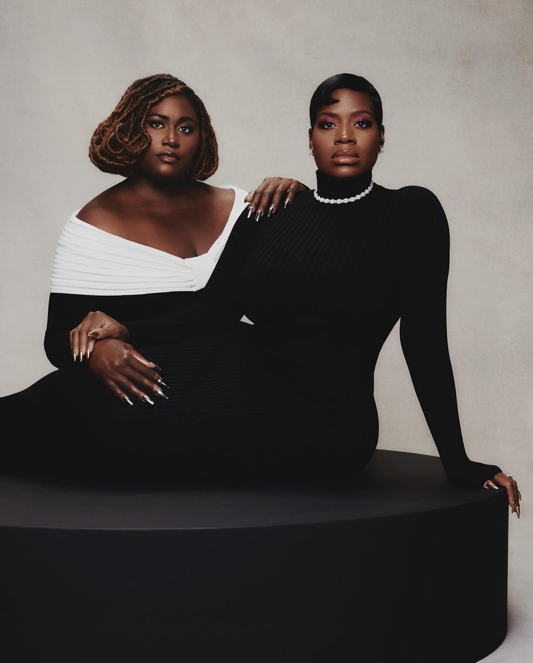Two adult Black women in elegant dresses pose in a portrait-style photo studio shot. One wears an off-the-shoulder white and black dress.