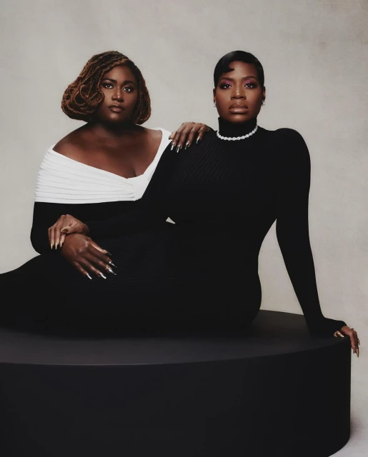 Two adult Black women in elegant dresses pose in a portrait-style photo studio shot. One wears an off-the-shoulder white and black dress.
