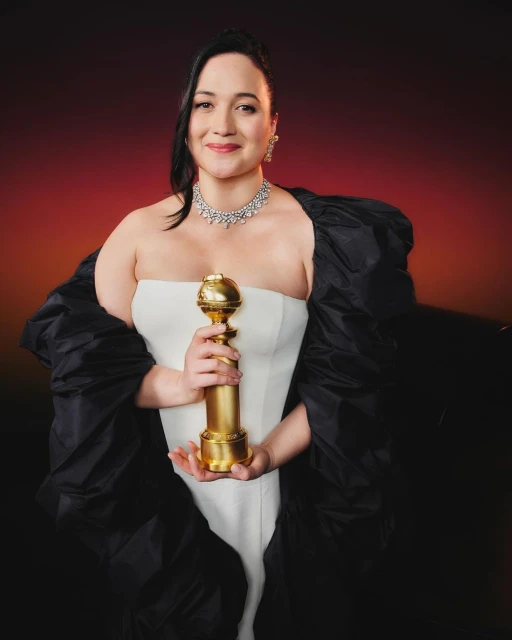 Adult woman holding a Golden Globe award. She is wearing a strapless white gown and a black shawl and diamond jewelry, studio lit.