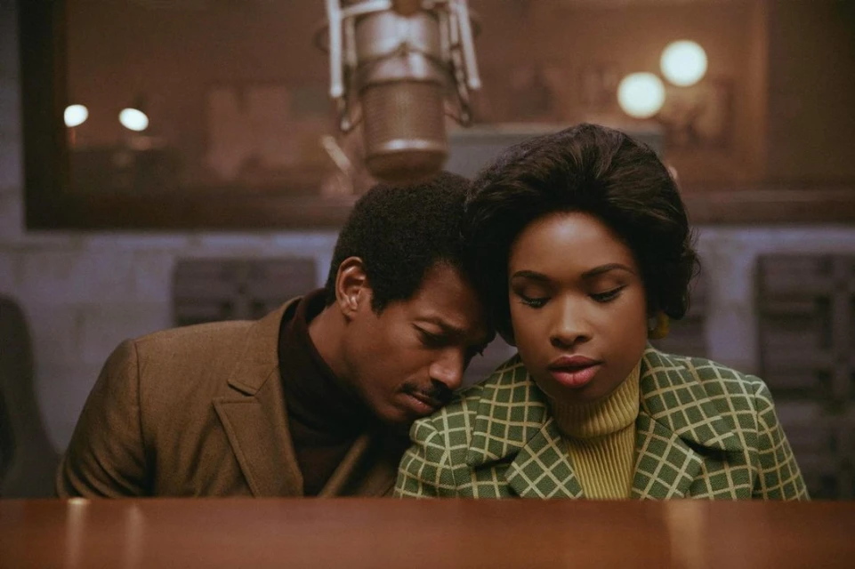 Two singers, an adult male and adult female, perform in a music studio. The woman sings with a microphone in front of her.