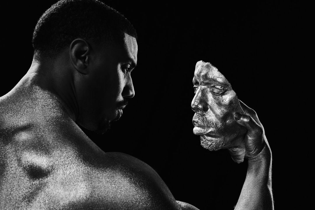 Monochrome portrait of Michael B. Jordan holding a metallic sculpted face mask against a black background.