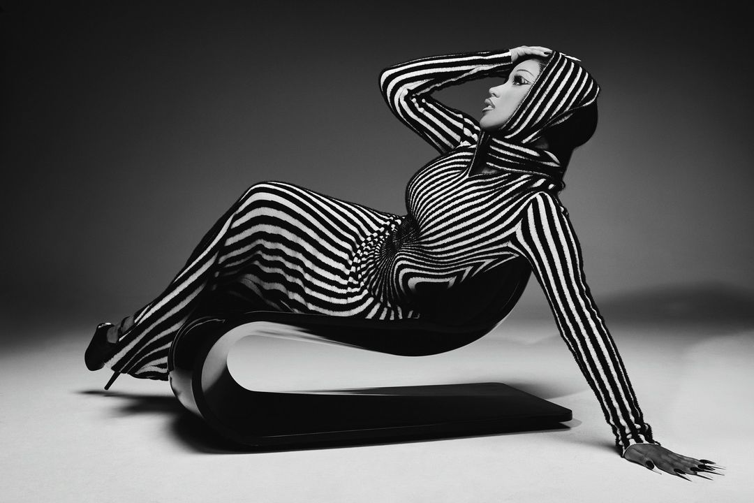 Striking black-and-white image of a woman in a striped hooded garment, reclining elegantly on a modern black chair, captured in a studio setting.