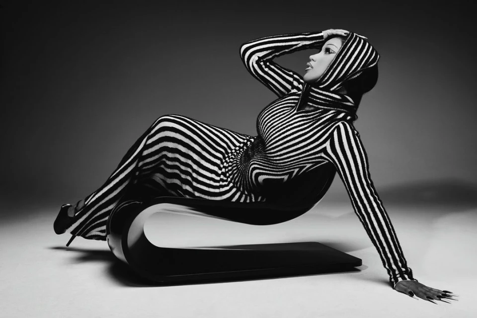 Striking black-and-white image of a woman in a striped hooded garment, reclining elegantly on a modern black chair, captured in a studio setting.