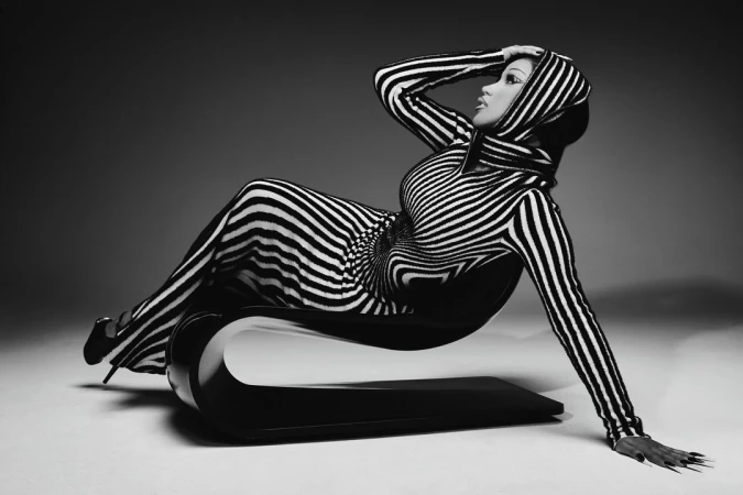 Striking black-and-white image of a woman in a striped hooded garment, reclining elegantly on a modern black chair, captured in a studio setting.