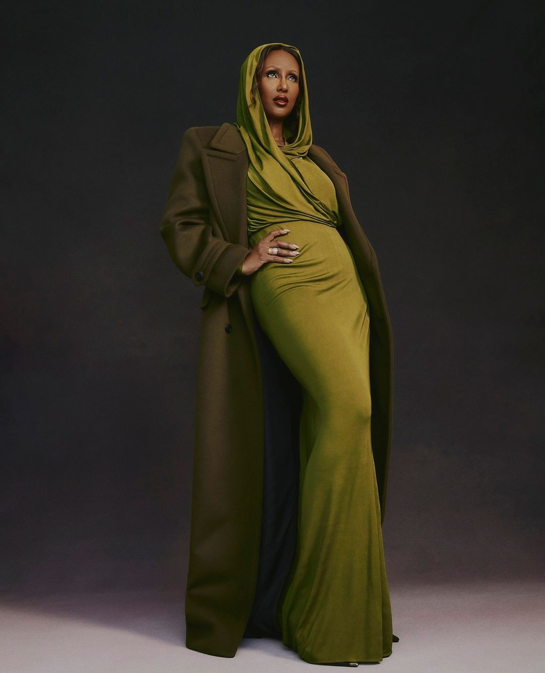 A woman in a long olive gown and matching coat poses in a studio with a dark background, creating a striking and elegant visual.
