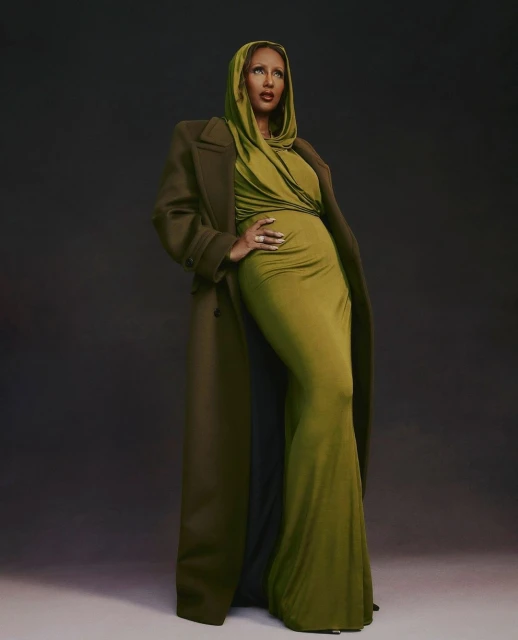 A woman in a long olive gown and matching coat poses in a studio with a dark background, creating a striking and elegant visual.