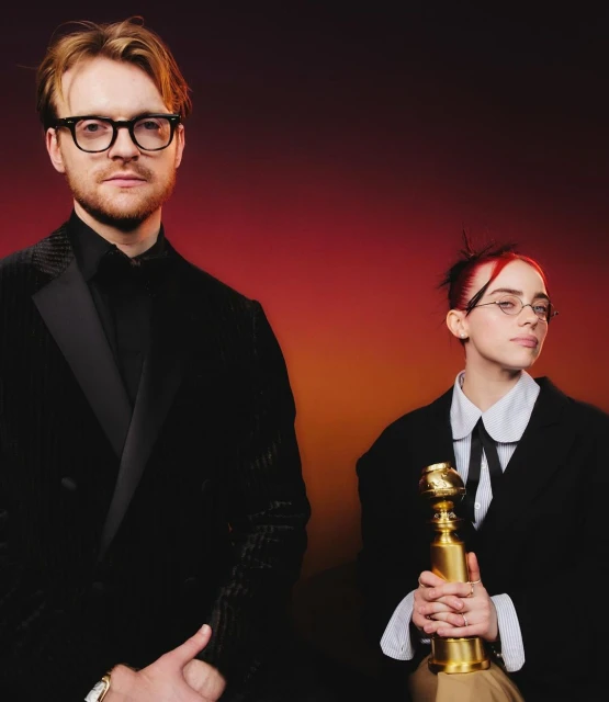 Finneas O'Connell and Billie Eilish stand indoors with a Golden Globe award, both in dark suits against a gradient red background.