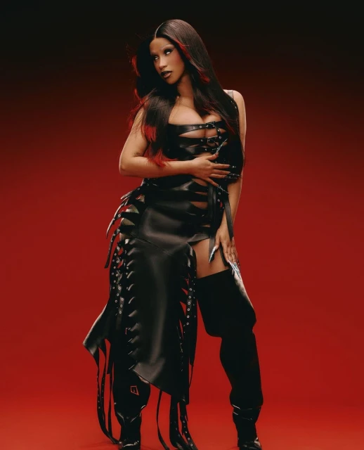A glamorous woman in a black leather outfit and thigh-high boots poses against a red backdrop, highlighting her edgy style and confidence.