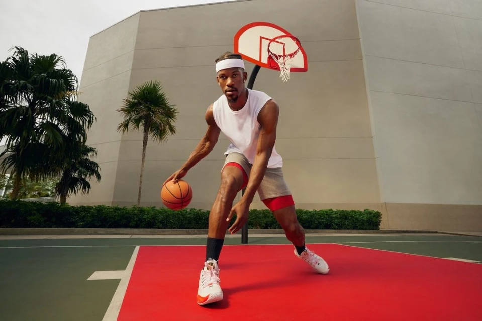 A basketball player dribbles a basketball on an outdoor court, wearing a white sleeveless shirt and grey shorts, with trees and a building in the background.